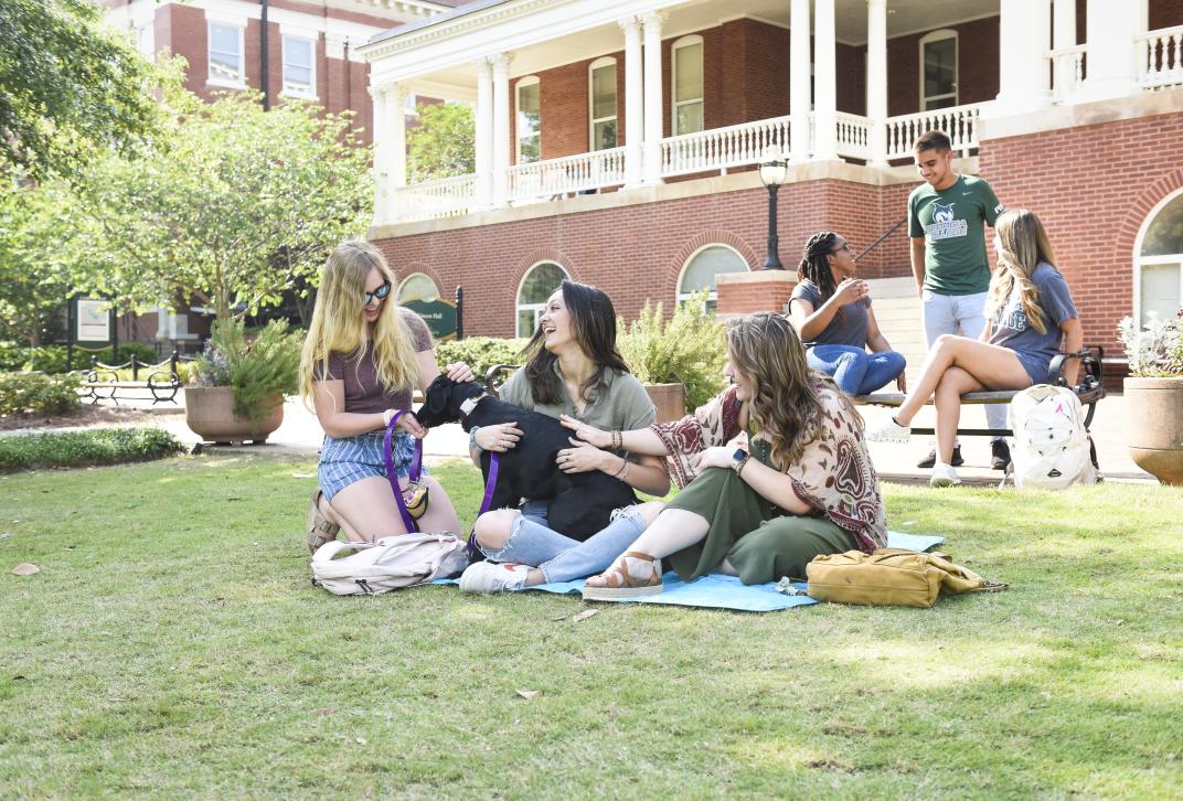 Students sit on front campus with a dog and enjoy the sunshine.