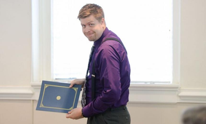 White man in purple shirt, short hair, holds certificate, turns, smiles.