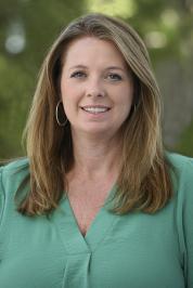Outdoor professional headshot of Amy Whatley, smiling with shoulder length blonde hair and hoop earrings, wearing a green v-neck blouse, against a softly blurred green background of foliage. 