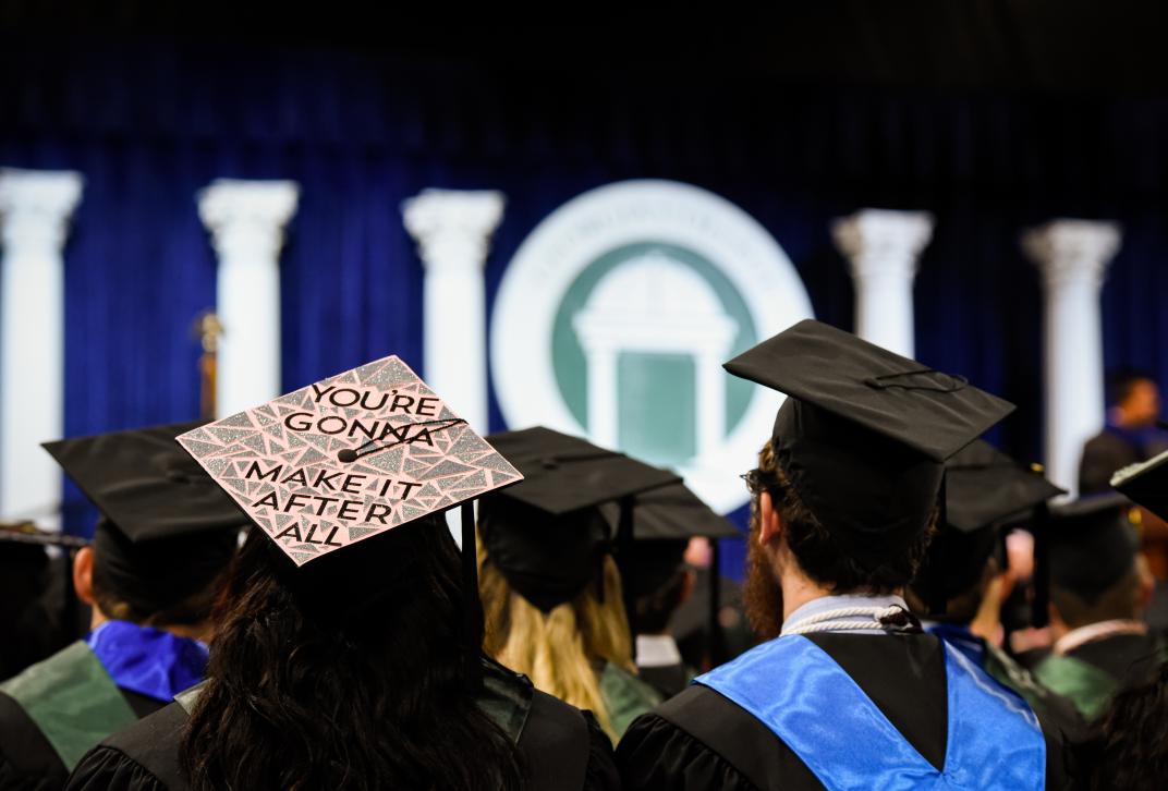 Picture of students facing the stage during commencement.  One cap is decorated with the phrase "you're gonna make it after all."