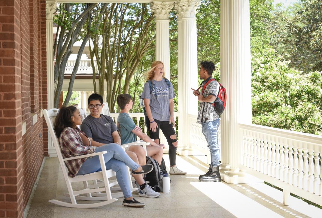 A group of five students is pictured on the front porch of Atkinson Hall. Several students sit in rocking chairs. They are all talking to each other.