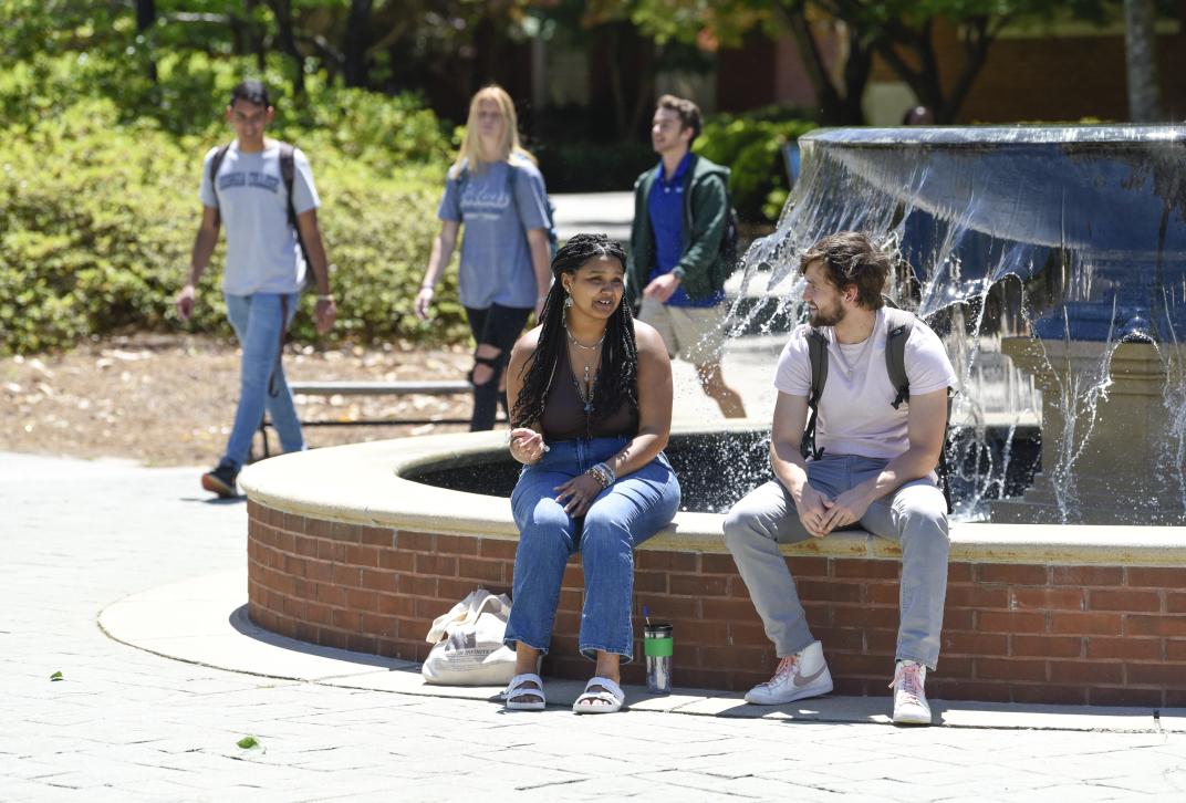 Two students sit by a fountain and talk. A group of students wearing Georgia College & State University t-shirts walk by in the background.