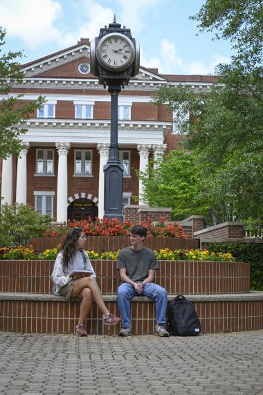 Two students sit on a ledge by the clock talking to each other