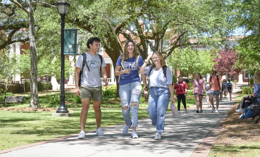 A group of students walk on the sidewalk next to front campus. They are smiling, laughing, and surrounded by large trees.